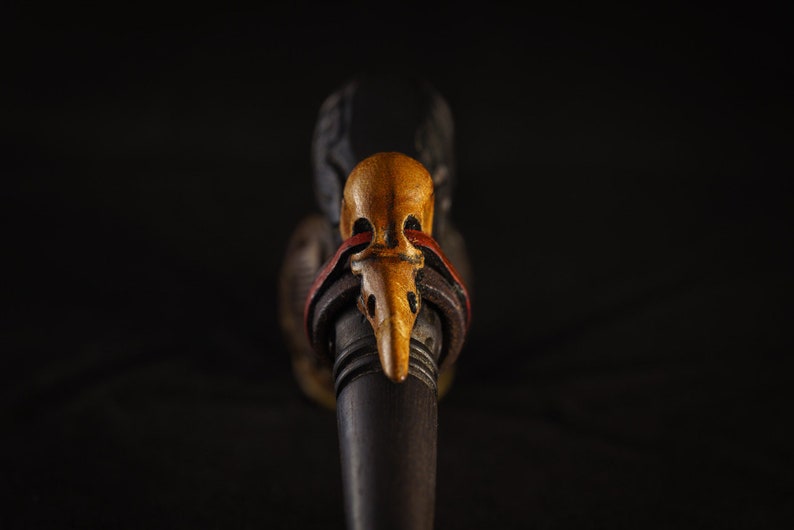 May include: A wooden pipe with a carved bird head design. The bird head is painted in shades of brown, yellow, and red. The pipe is decorated with a leather strap.