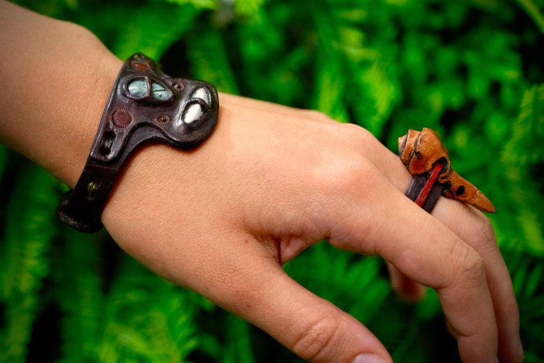 May include: A brown leather bracelet with a decorative metal design and a brown leather ring with a decorative metal design.