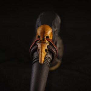 May include: A wooden pipe with a carved bird head mouthpiece. The bird head is painted in shades of yellow, orange, and red.