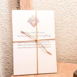 May include: A white card tied with twine, featuring text and a heart illustration. The text reads "The Surrender Novena: Let Jesus Take Care of Everything" and further details about the Novena. A religious item.