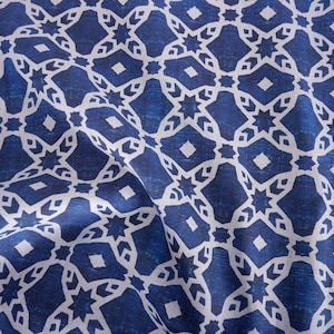 May include: Close-up of a blue and white patterned fabric. The design features a repeating geometric pattern of stars, diamonds, and other shapes. The fabric has a slightly textured appearance and is draped, creating folds and shadows.