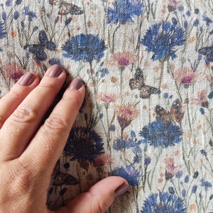 Könnte beinhalten: Nahaufnahme eines beigefarbenen Stoffes mit einem sich wiederholenden Muster aus blauen Blumen und braunen Schmetterlingen. Der Stoff hat eine strukturierte Leinenoptik.