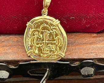 Op de afbeelding: Een gouden hanger in de vorm van een munt met een kruisontwerp en een lus aan de bovenkant. De hanger ligt op een houten oppervlak.