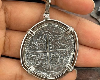 Op de afbeelding: Zilveren hanger met een replica van een Spaanse koloniale munt met een kruis en een wapenschild. De munt is ingelijst in een zilveren frame met decoratieve accenten.