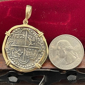 Può includere: Un ciondolo placcato oro con una moneta spagnola d'argento con un designo di stemma. La moneta è circondata da una cornice dorata e ha un anello in alto per appenderla. Il ciondolo è mostrato accanto a una moneta da 20 centesimi di euro per il confronto delle dimensioni.