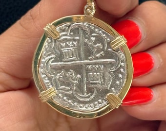 Op de afbeelding: Een zilveren munt hanger met een gouden frame. De munt heeft een kruis met vier schilden en een halve maan. De hanger wordt vastgehouden in een hand met rode nagellak.
