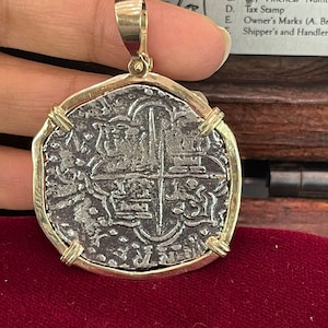 Puede incluir: Una moneda de plata con un colgante de oro, que presenta una cruz y otros diseños intrincados. La moneda está montada sobre un fondo de terciopelo rojo.