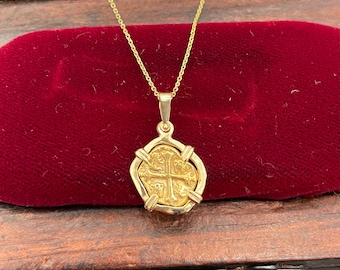 Op de afbeelding: Een gouden munt ketting met een kruisvormig hanger. De munt is ingelijst in een gouden frame en hangt aan een delicate gouden ketting. De ketting wordt gepresenteerd in een houten doos met een rode fluwelen voering.