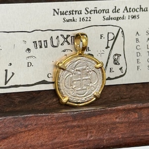 Puede incluir: Una moneda de plata con un marco de oro, que presenta una cruz y la inscripción "UXI". La moneda está sobre una tarjeta blanca con el texto "Nuestra Señora de Atocha" y las fechas "Sunk: 1622" y "Salvaged: 1985".