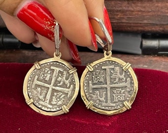Könnte beinhalten: Ein Paar silberne Ohrringe mit goldenen Akzenten. Jeder Ohrring zeigt eine silberne Münze mit einem Kreuz und einem Löwenmotiv. Die Münzen sind von einem goldenen Rand umgeben.