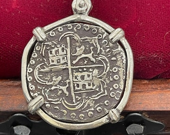 May include: A silver pendant featuring a replica of a Spanish eight reales coin, also known as a piece of eight. The coin features a coat of arms with a castle and a lion. The pendant is on a wooden surface.