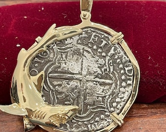 May include: A gold pendant featuring a shark biting into a silver coin. The coin has the inscription "REX ET INDIAE" and a cross design.