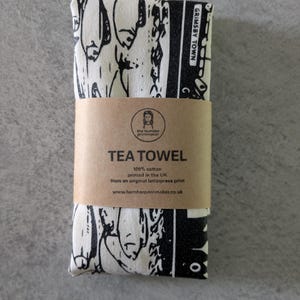 May include: A folded off-white tea towel featuring a black graphic print of fish and the text Grimsby Town. A brown paper band wraps around the centre, printed with the text TEA TOWEL, 100% cotton, printed in the UK, and a website address.
