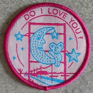 Op de afbeelding: Een geborduurde patch in roze en wit met een halve maan, een ster en de tekst "Do I Love You?"