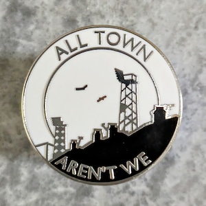 Può includere: Spilla rotonda in smalto con bordo argentato. La spilla presenta una silhouette di paesaggio urbano in bianco e nero con le parole "ALL TOWN AREN'T WE" in lettere argentate. Il design include una torre radio e uccelli.
