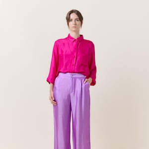 Può includere: Una donna indossa una camicia rosa acceso con bottoni e pantaloni larghi color lilla. È in piedi con le mani in tasca.