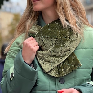 May include: A quilted olive green velvet scarf with white stitching is worn over a green jacket. The scarf is wrapped around the neck. A red and green bag is held in the hand.