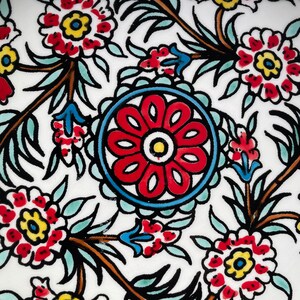 Puede incluir: Un azulejo de cer&aacute;mica blanco con un patr&oacute;n floral rojo, azul y amarillo. El azulejo presenta una gran flor estilizada en el centro rodeada de flores y hojas m&aacute;s peque&ntilde;as.