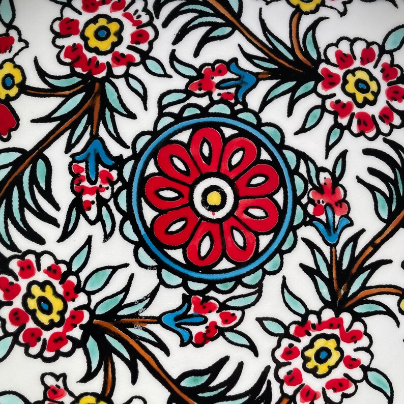 Puede incluir: Un azulejo de cer&aacute;mica blanco con un patr&oacute;n floral rojo, azul y amarillo. El azulejo presenta una gran flor estilizada en el centro rodeada de flores y hojas m&aacute;s peque&ntilde;as.