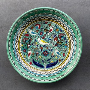 Op de afbeelding: Een groene keramische kom met een wit en blauw bloemenontwerp en kleurrijke vogels. De kom heeft een gegolfde rand en een witte rand.