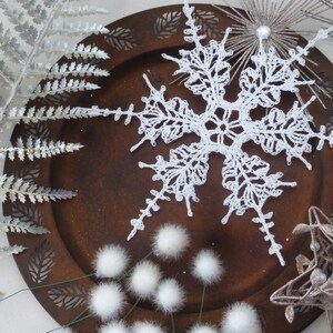 May include: A white crocheted snowflake ornament on a brown wooden plate with white fluffy pom-poms and silver fern sprigs.