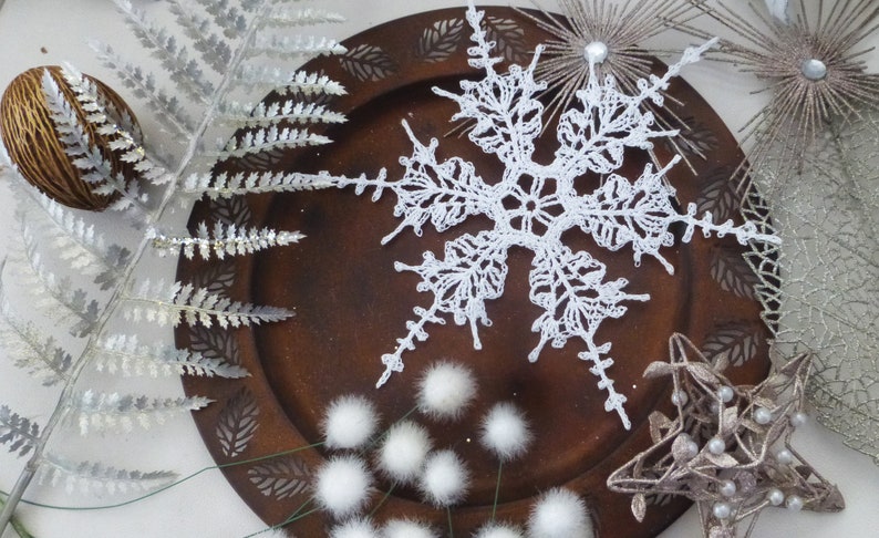 May include: A white crocheted snowflake ornament on a brown wooden plate with white fluffy pom-poms and silver fern sprigs.