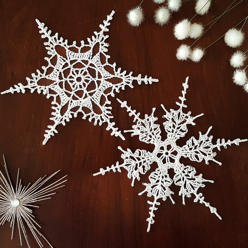 May include: Two white crocheted snowflakes on a brown wooden surface. The snowflakes are delicate and intricate, with a six-pointed star shape.