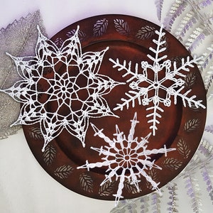 Op de afbeelding: Drie witte gehaakte sneeuwvlokken op een bruin houten bord met een decoratief ontwerp. De sneeuwvlokken zijn van verschillende grootte en hebben ingewikkelde details.