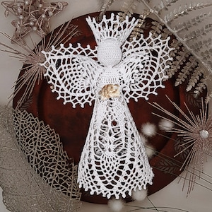 May include: A handmade white crochet angel with outstretched wings and a halo. The angel has a gold-colored accent on its chest and is displayed on a dark brown wooden plate, surrounded by decorative silver and gold accents.