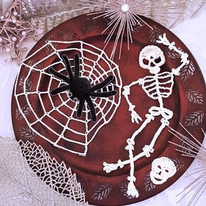 May include: A white crocheted skeleton with a spider web and a black spider on a red plate. The skeleton is posed with one arm raised and the other arm bent at the elbow. The spider web is in the background and the spider is in the center of the web.