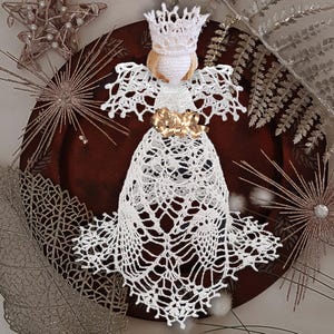 May include: A handmade white crochet angel ornament with a crown and gold ribbon. The angel has blonde hair and delicate, lacy wings and a skirt. The ornament is displayed on a dark brown plate with decorative accents.