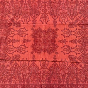 May include: A red textile with an intricate paisley pattern. The design features repeating floral and teardrop motifs in a darker shade of red, creating a layered effect. A white border with a delicate pattern frames the edges.