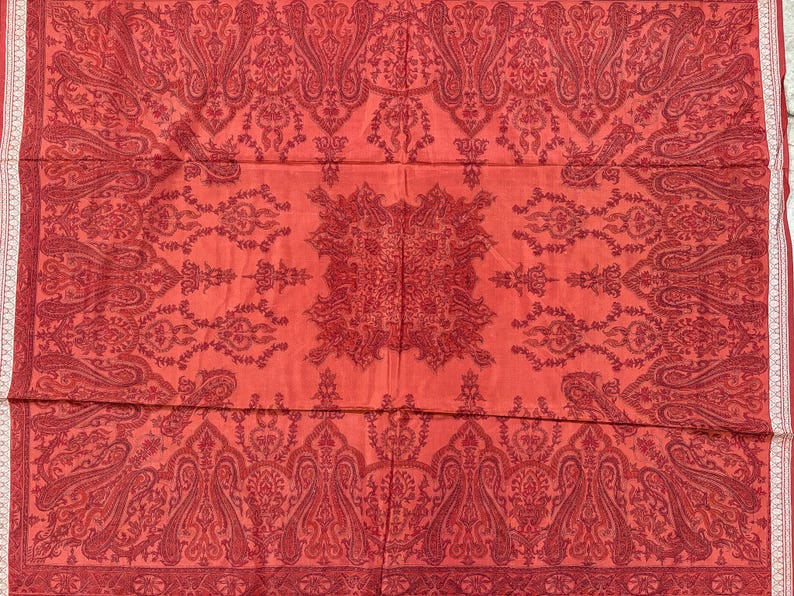 May include: A red textile with an intricate paisley pattern. The design features repeating floral and teardrop motifs in a darker shade of red, creating a layered effect. A white border with a delicate pattern frames the edges.