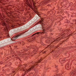 May include: A folded textile with a paisley pattern in shades of red and orange. The fabric has a detailed design with floral and curved elements. The border features a white and silver geometric pattern.