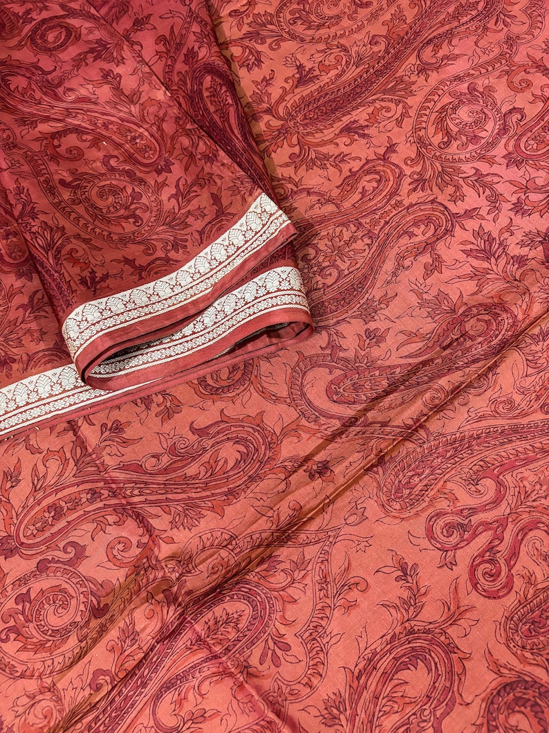 May include: A folded textile with a paisley pattern in shades of red and orange. The fabric has a detailed design with floral and curved elements. The border features a white and silver geometric pattern.