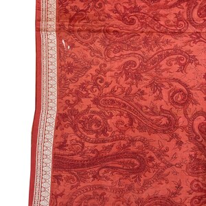 May include: A red paisley patterned fabric with a white and gold border. The fabric features a detailed design of swirling paisley shapes and floral elements. The border has a repeating geometric pattern.