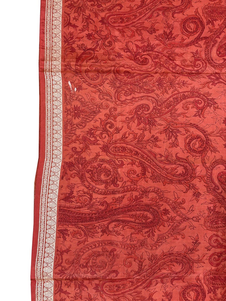May include: A red paisley patterned fabric with a white and gold border. The fabric features a detailed design of swirling paisley shapes and floral elements. The border has a repeating geometric pattern.