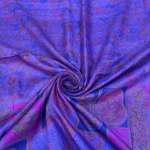 Op de afbeelding: Een wervelende stof in tinten paars, blauw en roze. Het textiel heeft een gedetailleerd bloemenpatroon met een mix van geometrische vormen. De textuur van de stof lijkt zacht en vloeiend, met een lichte glans.