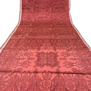 May include: A long, flowing textile in shades of red with an intricate paisley pattern. The fabric features a detailed design with repeating floral and teardrop motifs, creating a rich, textured appearance. A thin, white border runs along one edge.