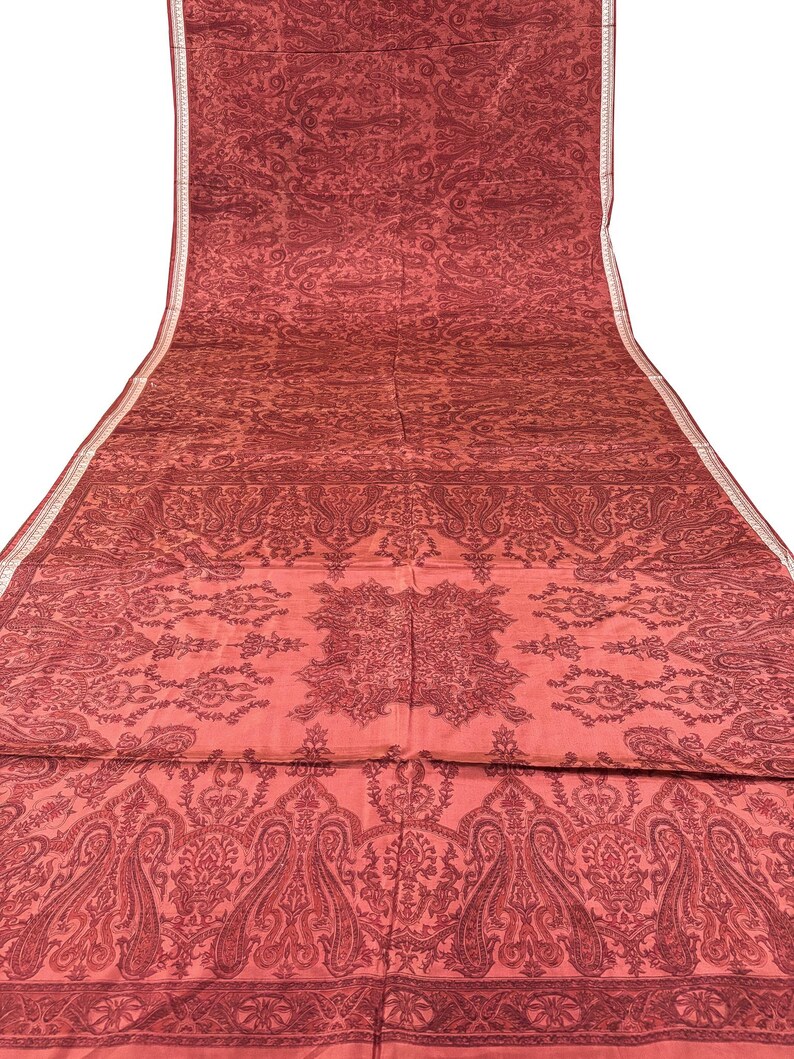 May include: A long, flowing textile in shades of red with an intricate paisley pattern. The fabric features a detailed design with repeating floral and teardrop motifs, creating a rich, textured appearance. A thin, white border runs along one edge.