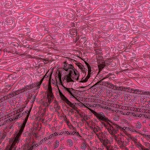 Könnte beinhalten: Ein lebendiger pinkfarbener Stoff mit einem komplizierten Paisley-Muster. Der Stoff hat einen Rand mit floralen und geometrischen Designs in Pink-, Lila- und Grautönen. Der Stoff ist drapiert und zeigt seine Textur und sein Design.