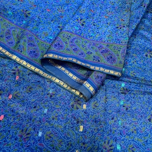 May include: A vibrant blue fabric with a detailed floral pattern and a decorative border. The border features paisley designs in green and purple, outlined with gold trim. The fabric has small, colorful accents throughout.