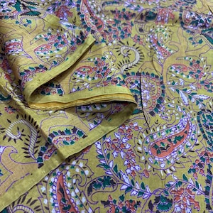 May include: A folded piece of fabric featuring a mustard yellow base color with an intricate paisley pattern. The design includes detailed motifs in shades of orange, green, purple, and white, creating a dense, repeating floral and teardrop print.