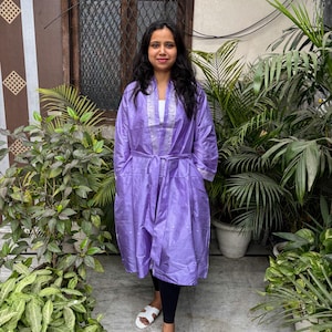 May include: A lilac-colored robe with a silver trim around the collar and cuffs. The robe has a matching belt and pockets. The person is wearing black leggings and white sandals. The background includes lush green plants and a window.