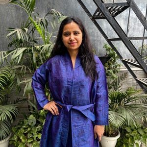 May include: A long, flowing robe in shades of blue and purple, with a matching belt tied at the waist. The robe has a subtle pattern and is made of a silky material. The person is standing in front of a backdrop of plants and a metal staircase.
