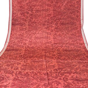 May include: A large, red textile with an all-over paisley pattern. The fabric has a detailed design in a darker shade of red, with a white border along one edge. The textile is likely a decorative fabric or a garment.