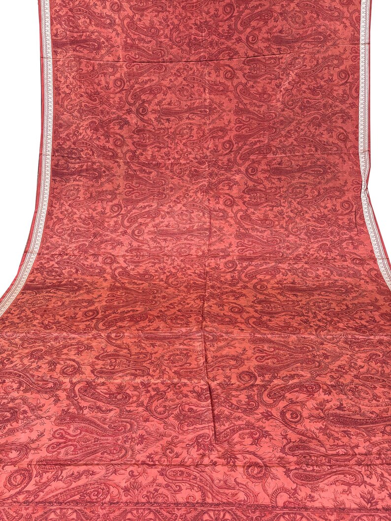 May include: A large, red textile with an all-over paisley pattern. The fabric has a detailed design in a darker shade of red, with a white border along one edge. The textile is likely a decorative fabric or a garment.
