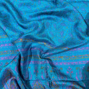 Op de afbeelding: Een levendige turquoise stof met een paisley patroon in paarse en gouden tinten. De stof heeft een zijdeachtige glans en is gedrapeerd, wat de textuur en de vloeiendheid van het materiaal laat zien. Het ontwerp bevat gedetailleerde bloemen- en druppelmotieven.