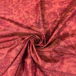 May include: Close-up of a vibrant, flowing fabric with a paisley pattern. The fabric is a blend of coral and deep red hues, with intricate floral and swirling designs. The material appears soft and luxurious, with visible folds and textures.