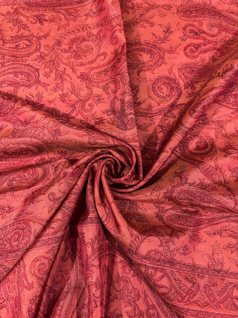 May include: Close-up of a vibrant, flowing fabric with a paisley pattern. The fabric is a blend of coral and deep red hues, with intricate floral and swirling designs. The material appears soft and luxurious, with visible folds and textures.